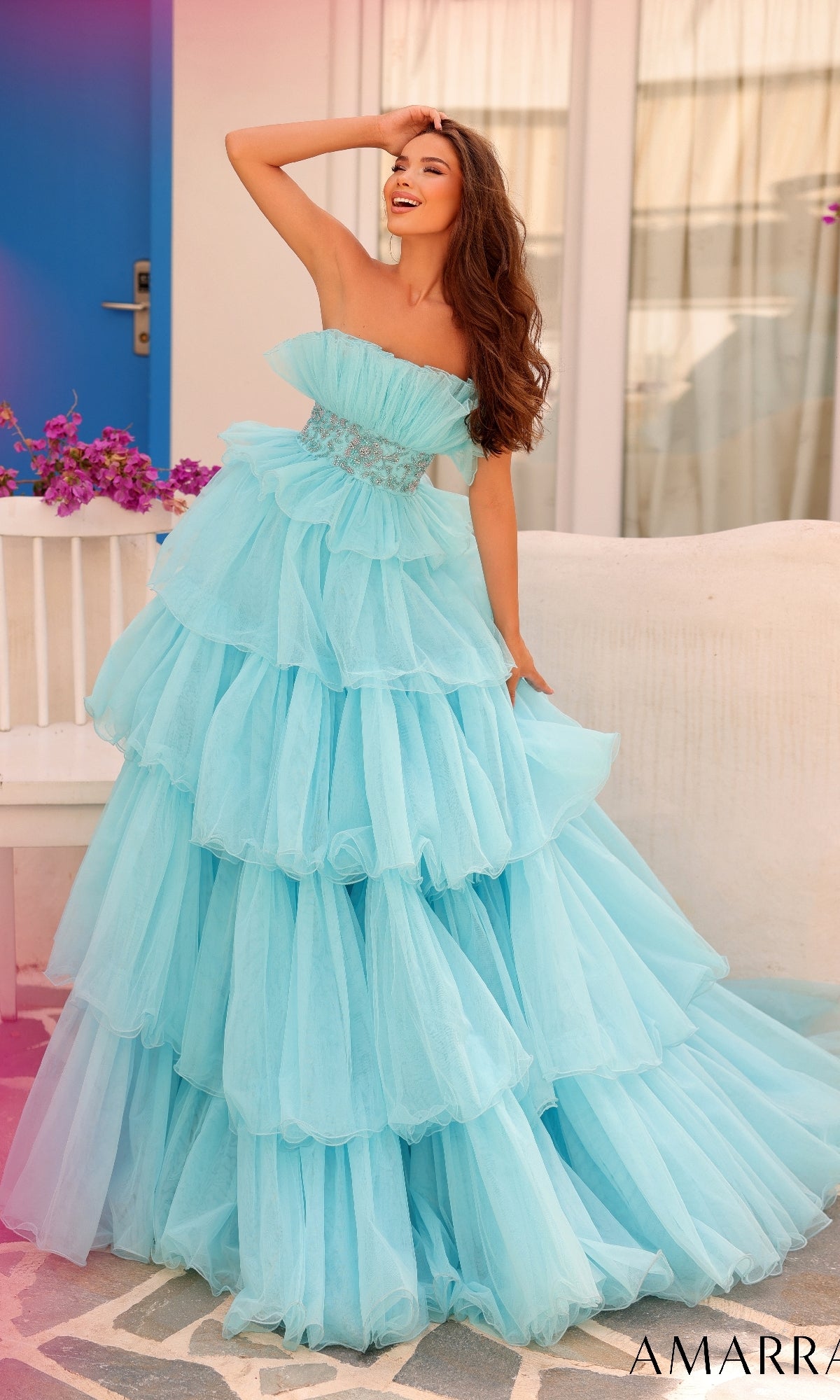 A woman poses outdoors by a white bench and blue door, smiling with one hand on her head, while wearing the Long Formal Dress 88083 by Amarra?a strapless gown featuring a beaded waist and tiered light blue layered skirt.
